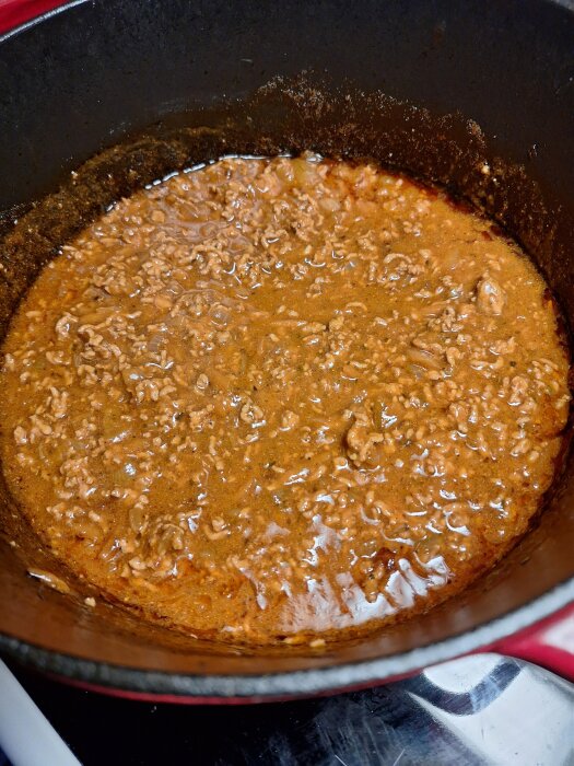 Köttfärssås kokar i en röd kastrull, med väl synlig vätska och en brun-orange färg på såsen.