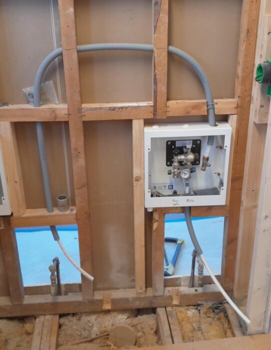Vägg under renovering med rör och kopplingsbox för tillopp och retur i radiatorkrets och golvvärmeslang, exponerad trästomme och isolering.