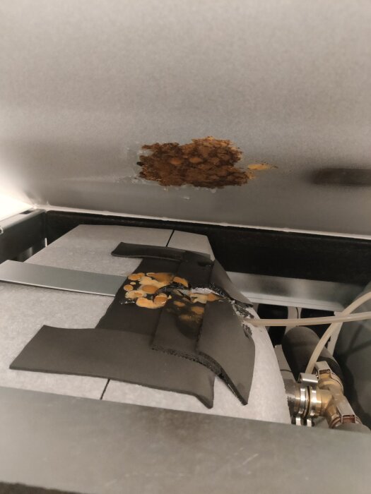 Rostfläck på insidan av en metallkåpa med skumisolering och rör.