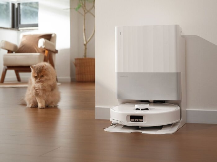 Robotdammsugare Roborock Qrevo Edge vid sin laddningsstation, med en katt i bakgrunden på ett trägolv.