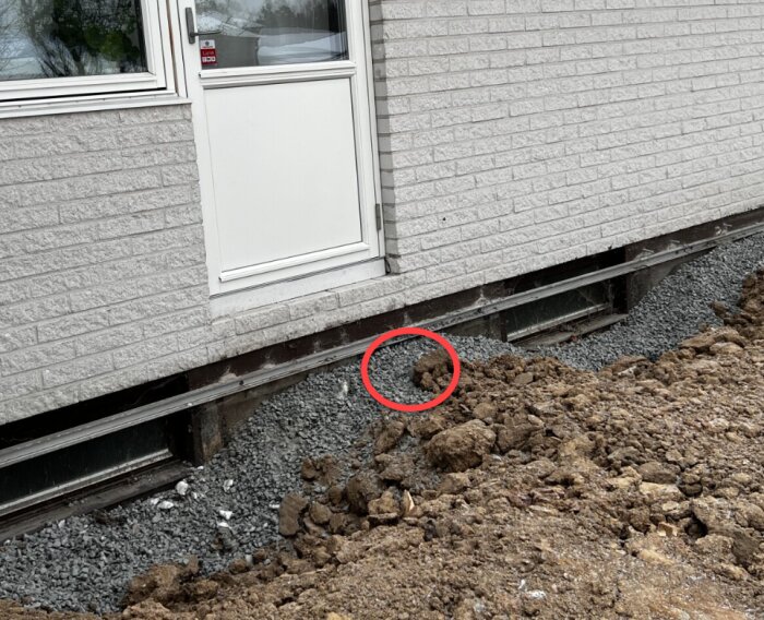 Fasad av ett hus med täckt ventil och grus vid grunden, samt en vit dörr ovanför marknivån.
