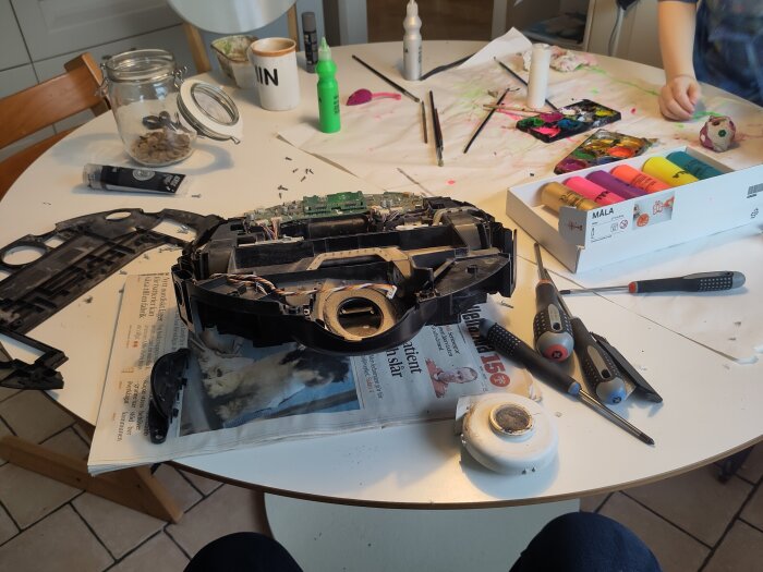 Öppen robotdammsugare på bord, omgiven av verktyg och målarfärg. Dammsugaren får service med nytt batteri, filter och borste.