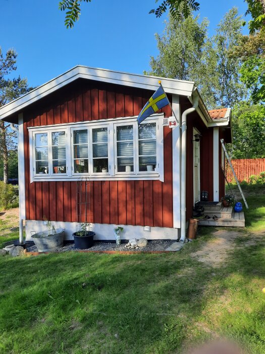 Rött hus med vita knutar, svensk flagga, och små trappsteg. Trädgård med gräs och blomkrukor i förgrunden. Klart väder och omgivande träd.