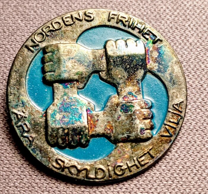 Traveled gamla märke med tre händer som greppar varandra, omgiven text: "Nordens frihet" och "Ära Skyldighet Vilja", mot turkos bakgrund.
