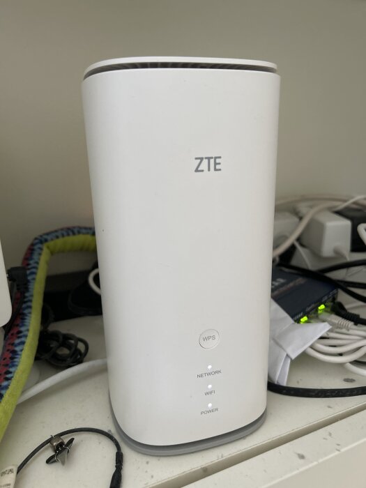 Vit ZTE MC888 Pro-router med lysande lampor för nätverk, WiFi och ström, omgiven av kablar och ett fodral av tyg i olika färger.