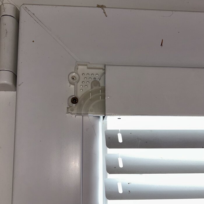 Nära bild på hörnet av en vit persienn där dragsnöret är borttaget, med synliga skruvar och fäste i ett fönster.