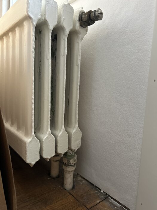En gammal vit radiator inomhus med rostiga ventiler och rör som går genom golvet.