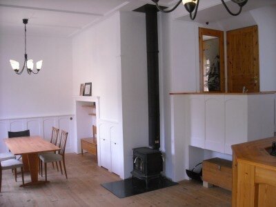 Vardagsrum med ljusa väggar, bröstpanel, en vedeldad kamin, träbord med stolar och taklampa i äldre stil.