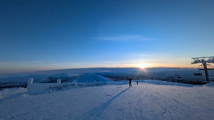 Solnedgång över snötäckt fjällandskap med två personer, en med skidor, och en skidlift till höger.