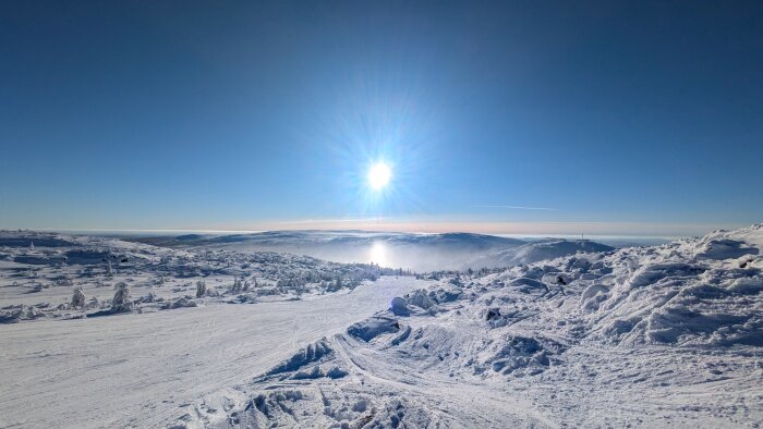Snötäckt bergslandskap med strålande sol på himlen, omgivande horisont och utsikt över vidsträckta fjäll.