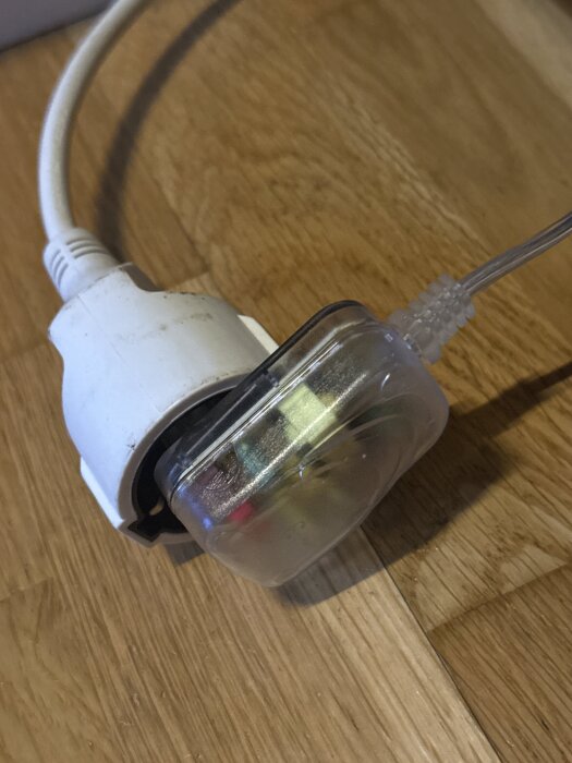 Genomskinlig elkontakt ansluten till en vit nätsladd på ett trägolv.