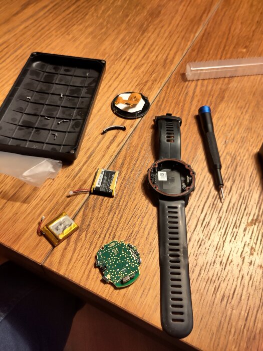 Delarna av en isärplockad smartwatch, inklusive batterier, skruvar och verktyg, ligger utspridda på ett träbord.
