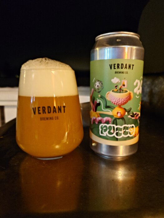 Ölglas och burk från Verdant Brewing Co med färgglad etikett, fyllt glas med skum på mörk bakgrund.