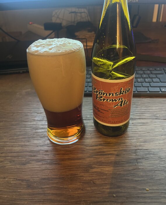 Ett glas fyllt med skummande Brännskär Brown Ale står bredvid en öppnad flaska på ett träbord framför ett tangentbord.