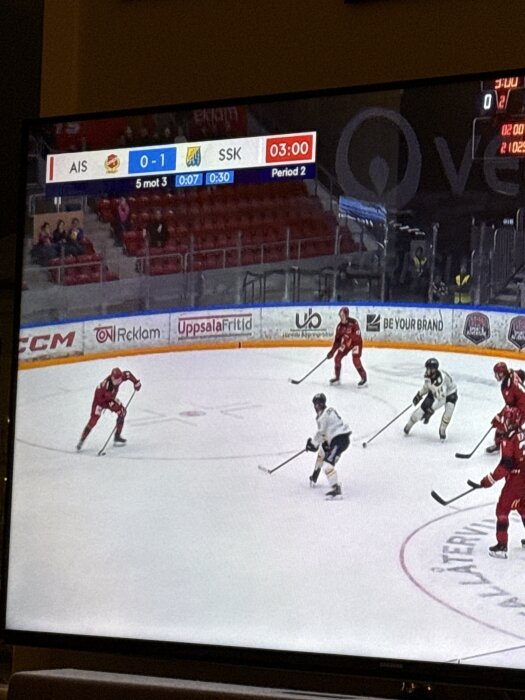 En hockeymatch visas på en TV, med spelare i rött och vitt på isen och poängtavlan visar ställningen 0-1 i andra perioden.