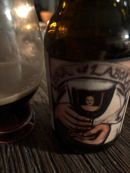 Flaska och glas med belgisk choklad stout, etikett visar händer som håller en bägare med en nunna. Träbord i bakgrunden, dämpad belysning.