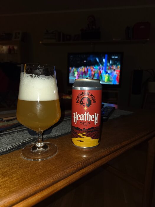 Ett glas öl och en burk Northern Monk Heathen Hazy IPA på ett bord framför en TV som visar ett färgglatt program.