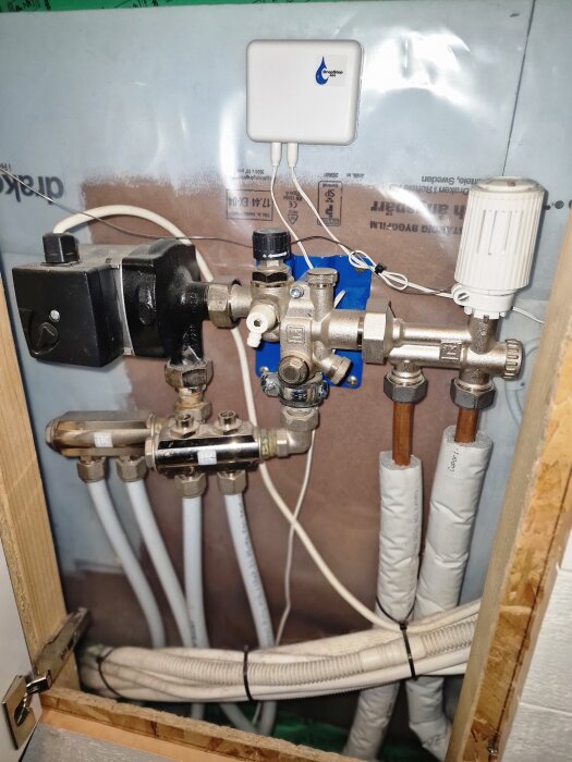 Bilden visar ett värmesystem med rör, kopplingar och en justeringsventil. Det finns en vit elektronisk modul kopplad med kablar.