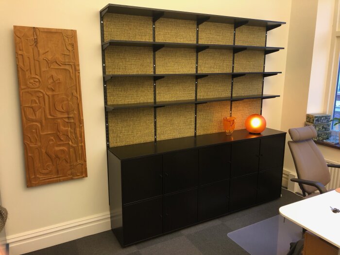 Svart skåp med hyllor mot beige vägg, orange lampa och reliefkonst av trä. Kontorsstol och skrivbord syns till höger.