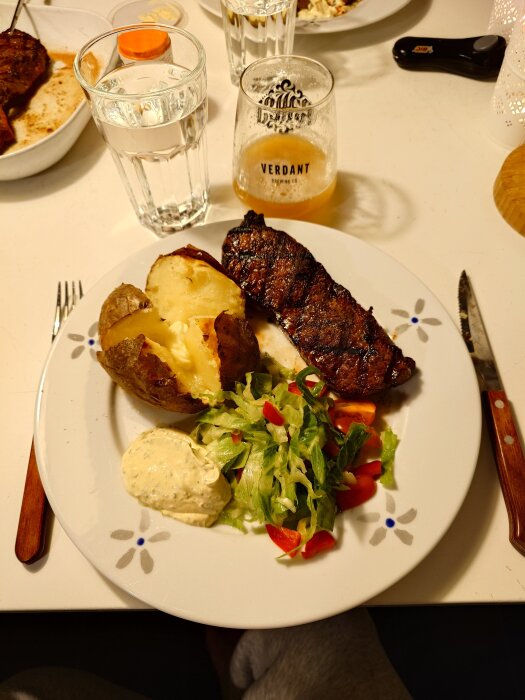 Grillad biff med bakad potatis, grönsallad och sås på tallrik. Glas med dryck och ett glas vatten bredvid.
