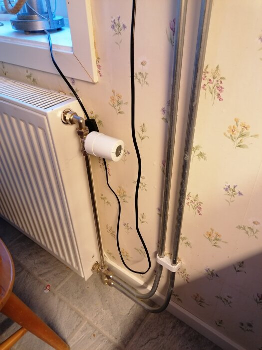 Värmeelement med rörkopplingar och termostatventil mot blommigt tapetserad vägg, sladd synlig vid sidan, del av golv och stolskant.