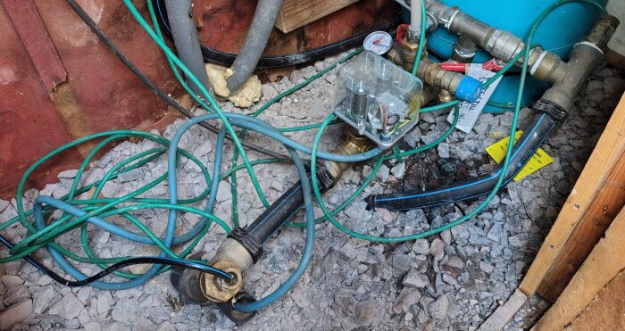 Vattenpumpinstallation med slangar och tryckmätare i en brunnsmiljö, grått grus, elektriska kopplingar. Problem med svagt vattenflöde.
