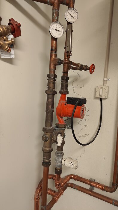 Rörsystem med ventiler och två termometrar monterade på kopparrör. En röd pumpenhet och en termostatventil syns i bilden.