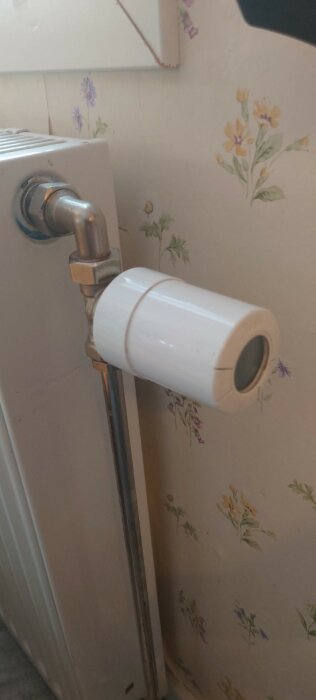 Vit termostatventil installerad på ett element, bakgrund med blommigt mönster på väggen.