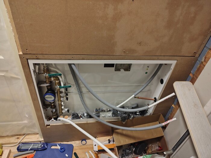 Installationsprocess av rör och fördelarskåp innanför en vägg, med exponerade vattenrör och verktyg på golvet.