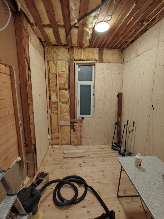 Rum under renovering med träväggar och isoleringsmaterial i taket. Ett fönster är synligt. Verktyg och dammsugare på golvet.