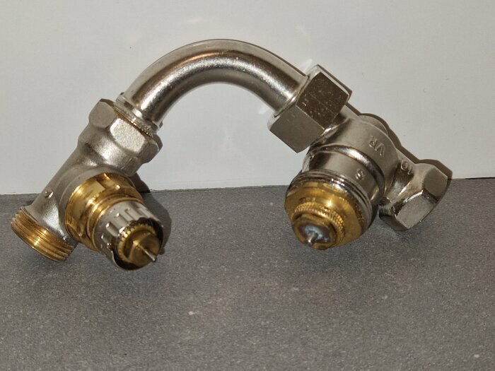 Två termostatventiler med urskruvade ventilinsatser och packboxar, en Danfoss och en MMA, ligger på en grå yta.