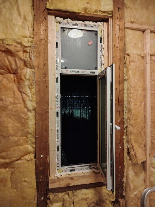 Ett fönster installerat i ett rum med isolering synlig runt om. Utsikt över en mörk skog genom det öppna fönstret.