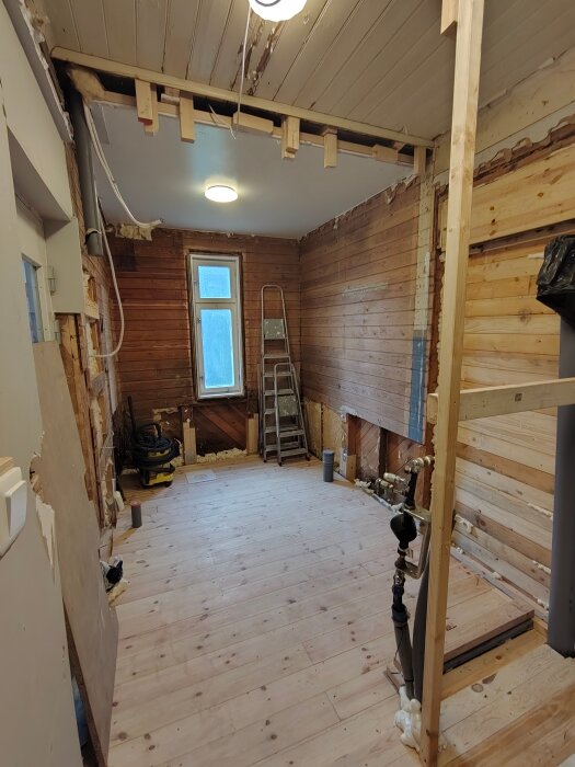 Rum under renovering med trägolv och öppnade väggar. Stege och verktygsslangar synliga, samt rörinstallation nära öppning i taket.
