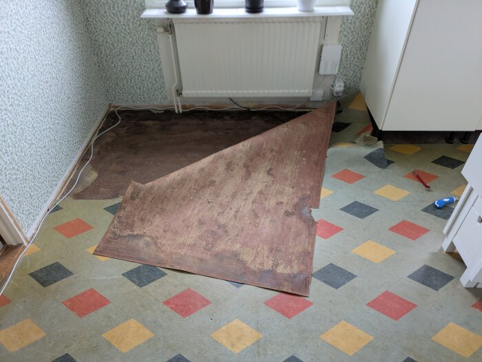 Golv från 60-talet med mönstrade färgglada rutor, delvis avtäckt. Rummet har vävda vita och gröna tapeter samt ett fönster med krukväxter.