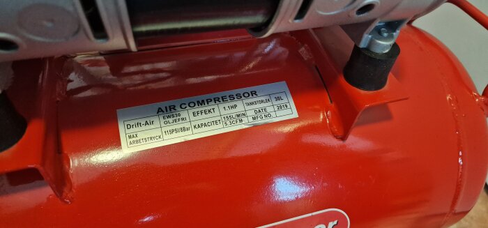 Röd luftkompressor med etikett: "Air Compressor, Effekt 1.1HP, Tankstorlek 30L, Max arbetstryck 115PSI/8bar, Kapacitet 155L/min, Datum 2019".