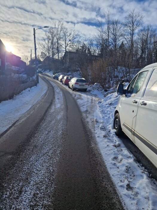 En smal, isig landsväg med parkerade bilar längs kanten, buskar och träd omgivna av snö, en vit skåpbil närmast i förgrunden.