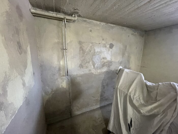 Tomt garageutrymme med målade betongväggar, synliga rör längs taket, och ett täckt föremål i hörnet.