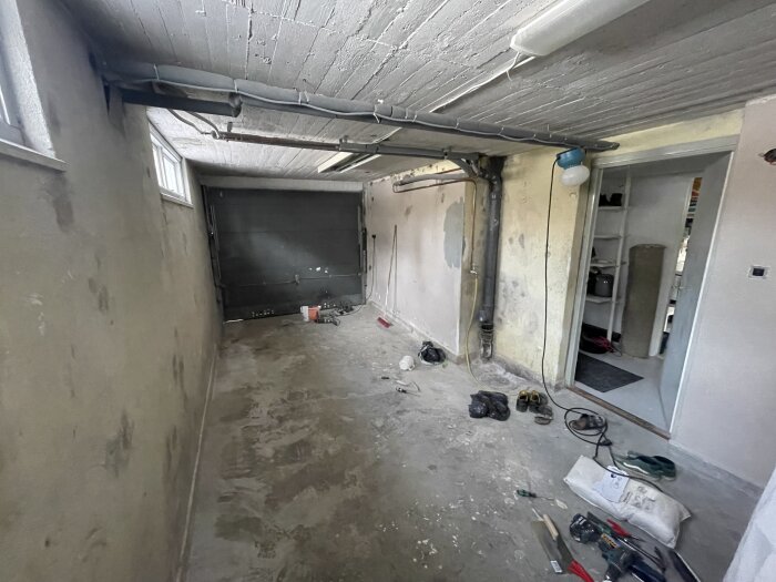 Smalt och tomt garage från 50-talet med framdragna rör och ett betonggolv. Dörr och hängande lampa syns, samt diverse verktyg på golvet.