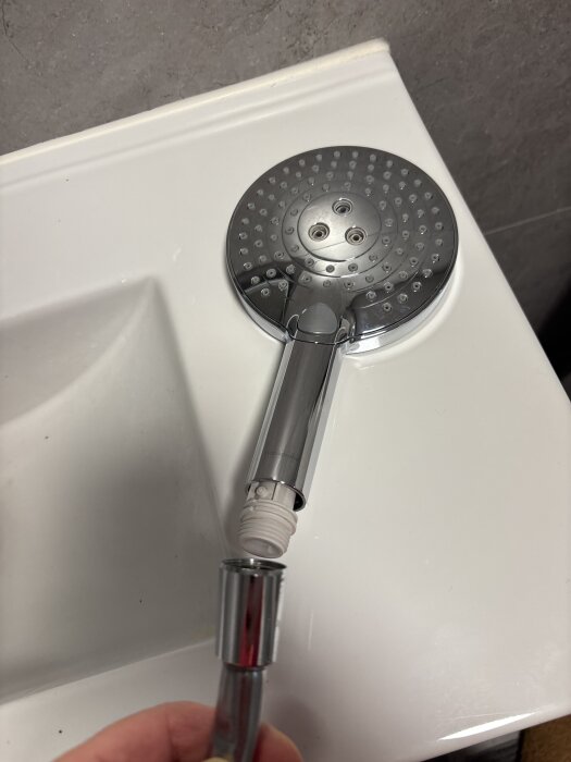 Ett duschmunstycke från Hansgrohe med en vit del fast i anslutningen, ligger på en vit badrumsyta.
