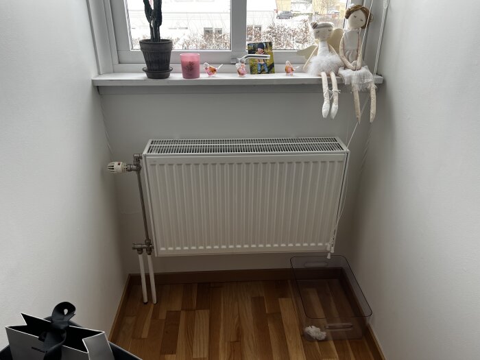 Element under ett fönster med leksaker på fönsterbrädan, kaktus och ljus ovanpå. Träparkettgolv. Plastbehållare på golvet till höger.
