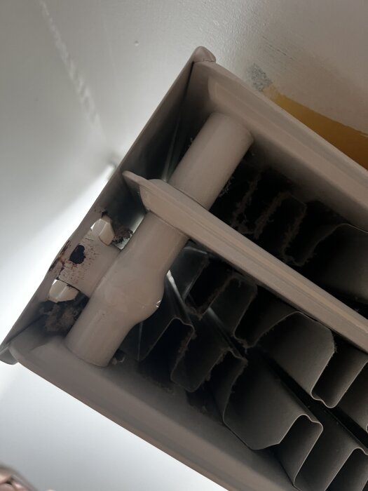 Närbild på en radiator med rostigt rör och damm mellan lamellerna, installerad nära en vägg med färgskador.