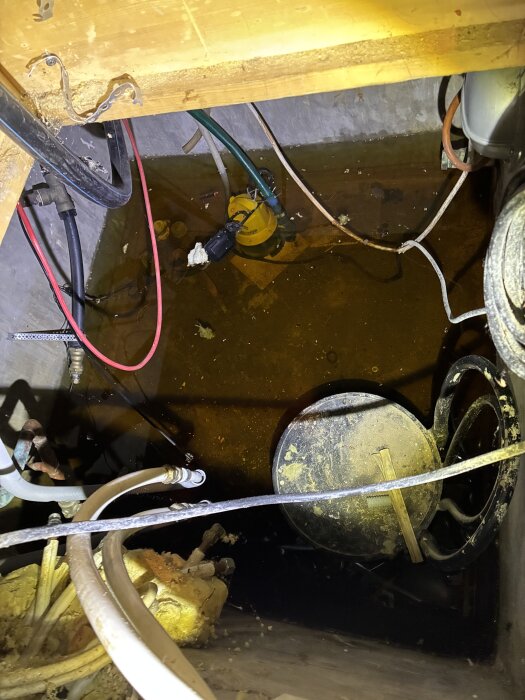 Utrymme under köket fyllt med vatten, kablar och en gul pump, omgiven av trä- och plastmaterial.