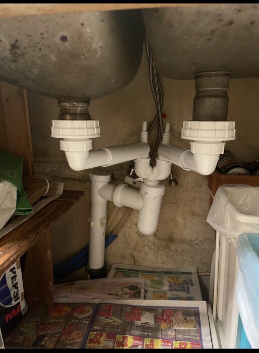 Undersida av ett dubbelt handfat med vit plastvattenlåsinstallation, synliga rör och tecken på läckage och stopp.