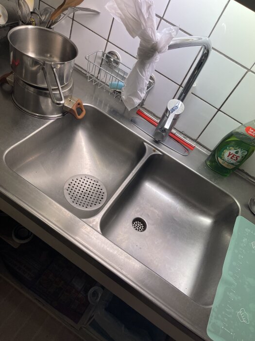 Dubbeldiskho i rostfritt stål med kastrull, diskmedel och knutna plastpåsar vid kranen. Betongvägg med kakel i bakgrunden.