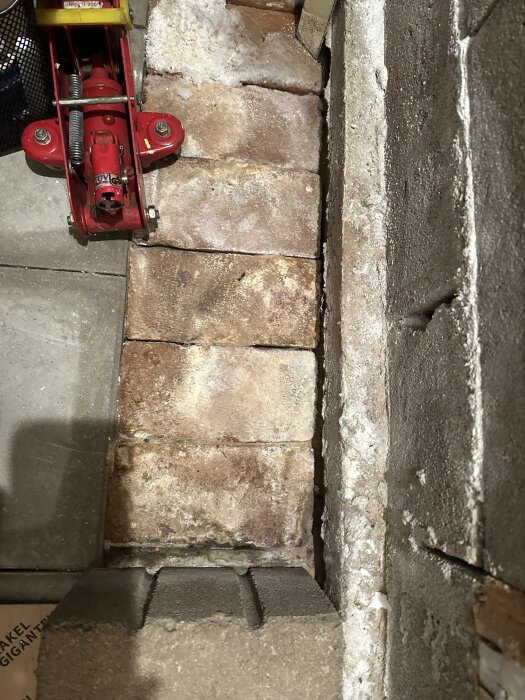Röd domkraft på betonggolv bredvid murade tegelstenar och cementvägg.