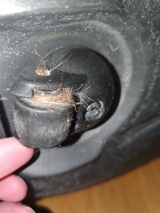 Närbild på en svart knapp med slitage och utspridda trådar eller fibrer. En finger vidrör knappen, bakgrunden är suddig.