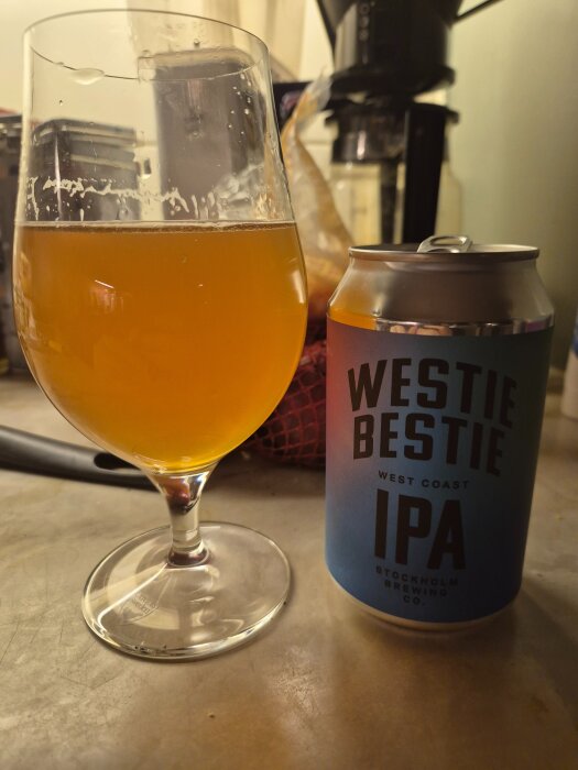 En ölburk med texten "Westie Bestie IPA" bredvid ett glas med halvt upphälld gyllene vätska, troligen öl, på ett köksbord.