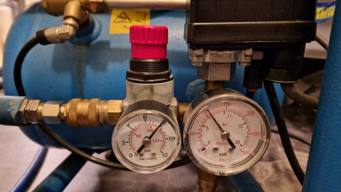 Eldriven kompressor med två tryckmätare och röd regulator, kopplad till en blå tank med slangar.