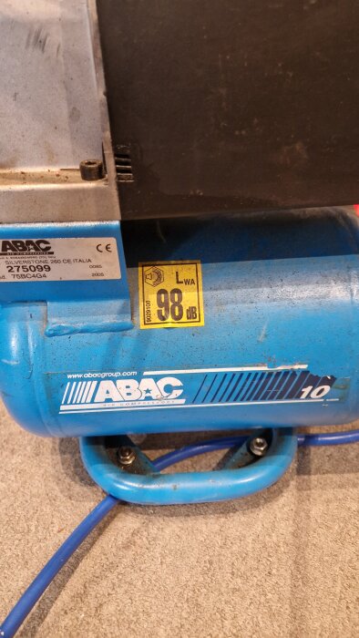 Blå kompressor från ABAC, modell Silverstone 260 CE, med ljudnivå på 98 dB. Märke på tanken och elkabel synlig på golvet.