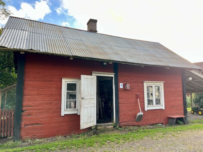 Rött timrat gårdshus med korrugerat tak, öppna dörrar och två fönster. En spade hänger på väggen, mark omgiven av gräs och grus.
